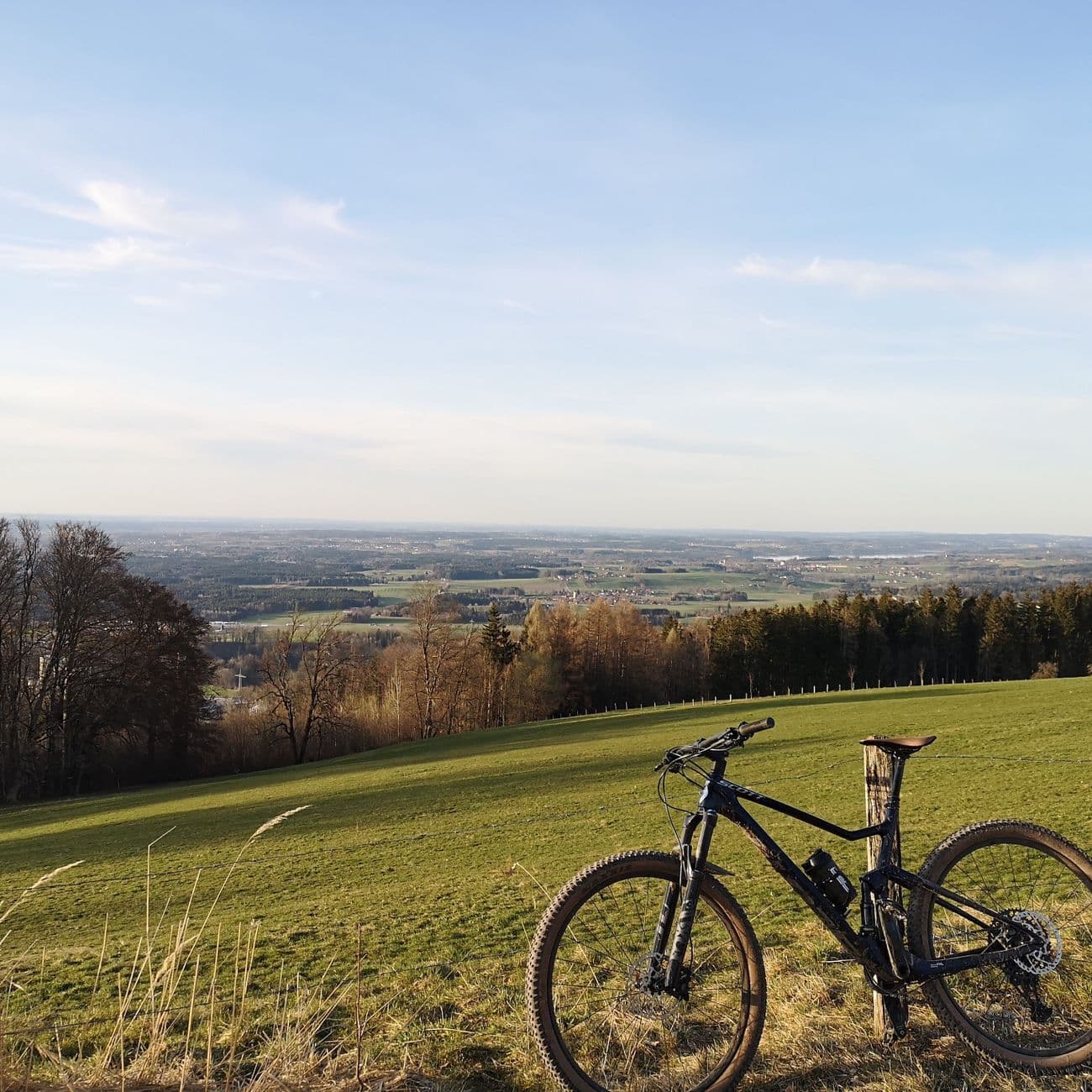 A dark blue Scott Spark mountain bike propped against a fence post on a grassy hillside, the Bavarian foothills fading to the horizon under a pale evening sky.