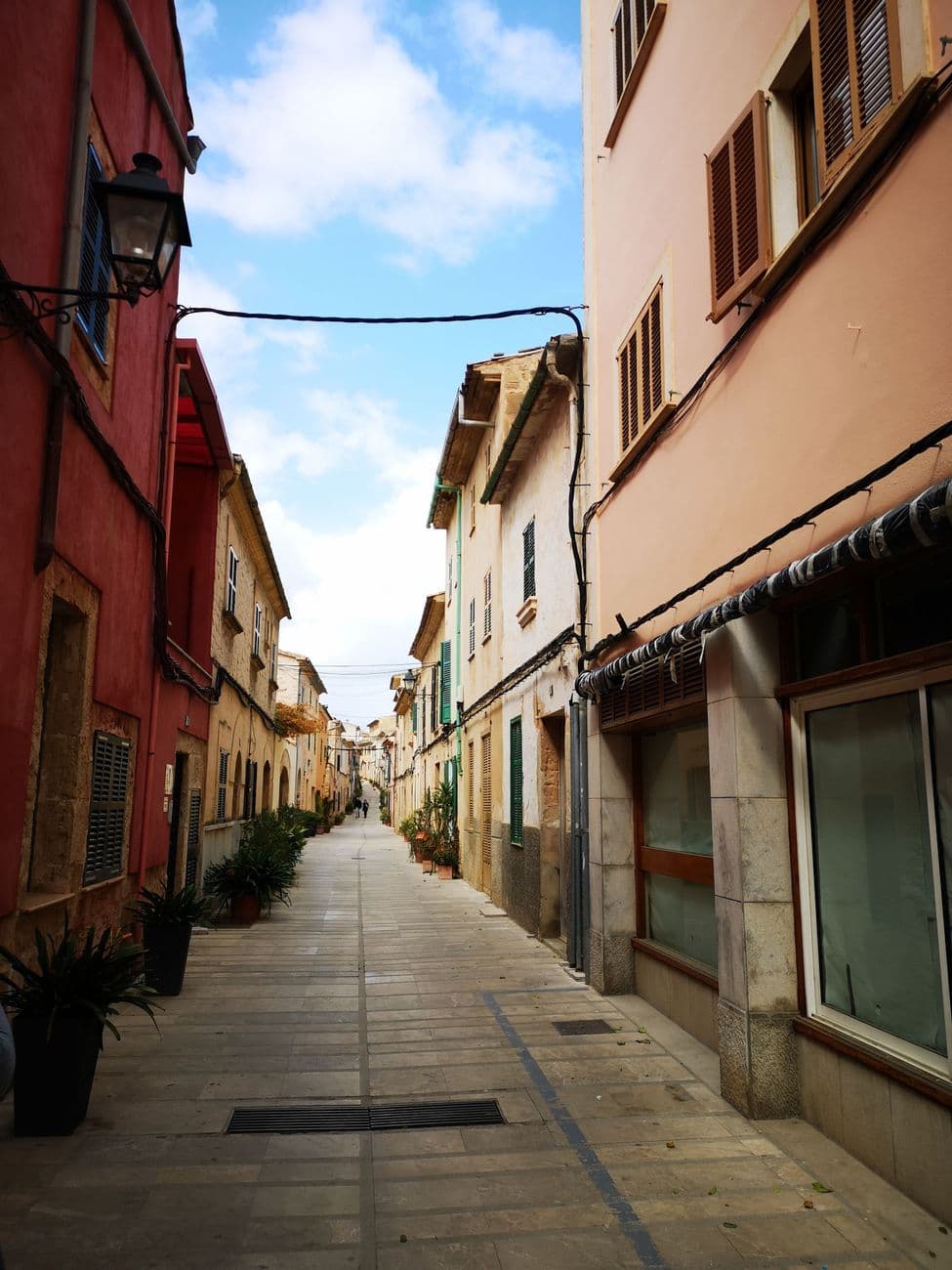 A narrow street in Alcúdia's old town, ochre and deep red housefronts with wooden shutters, potted plants along the paving.