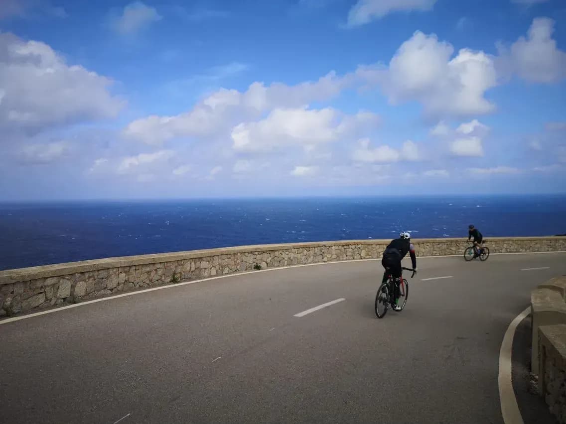 Two road cyclists on the coastal road at Cap Formentor, a low stone wall between them and the deep blue Mediterranean.