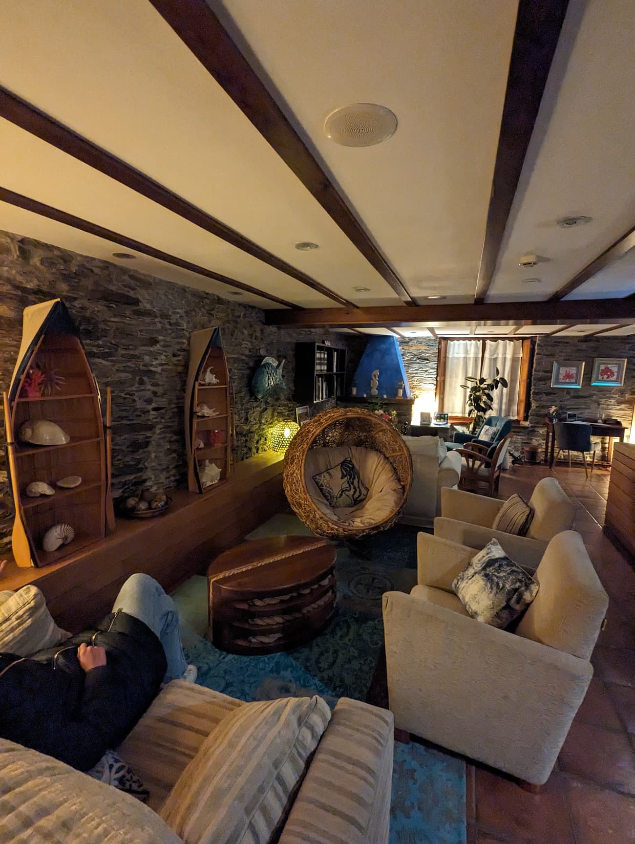 A stone-walled lounge lit warm at night: shelves built into upturned rowing boats hold shells, a hanging wicker chair sits on a blue patterned rug.