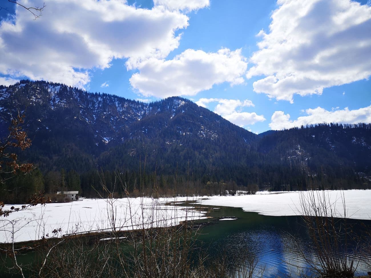 A partly thawed alpine lake, half still under snow, ringed by steep forested mountains under scattered cloud.