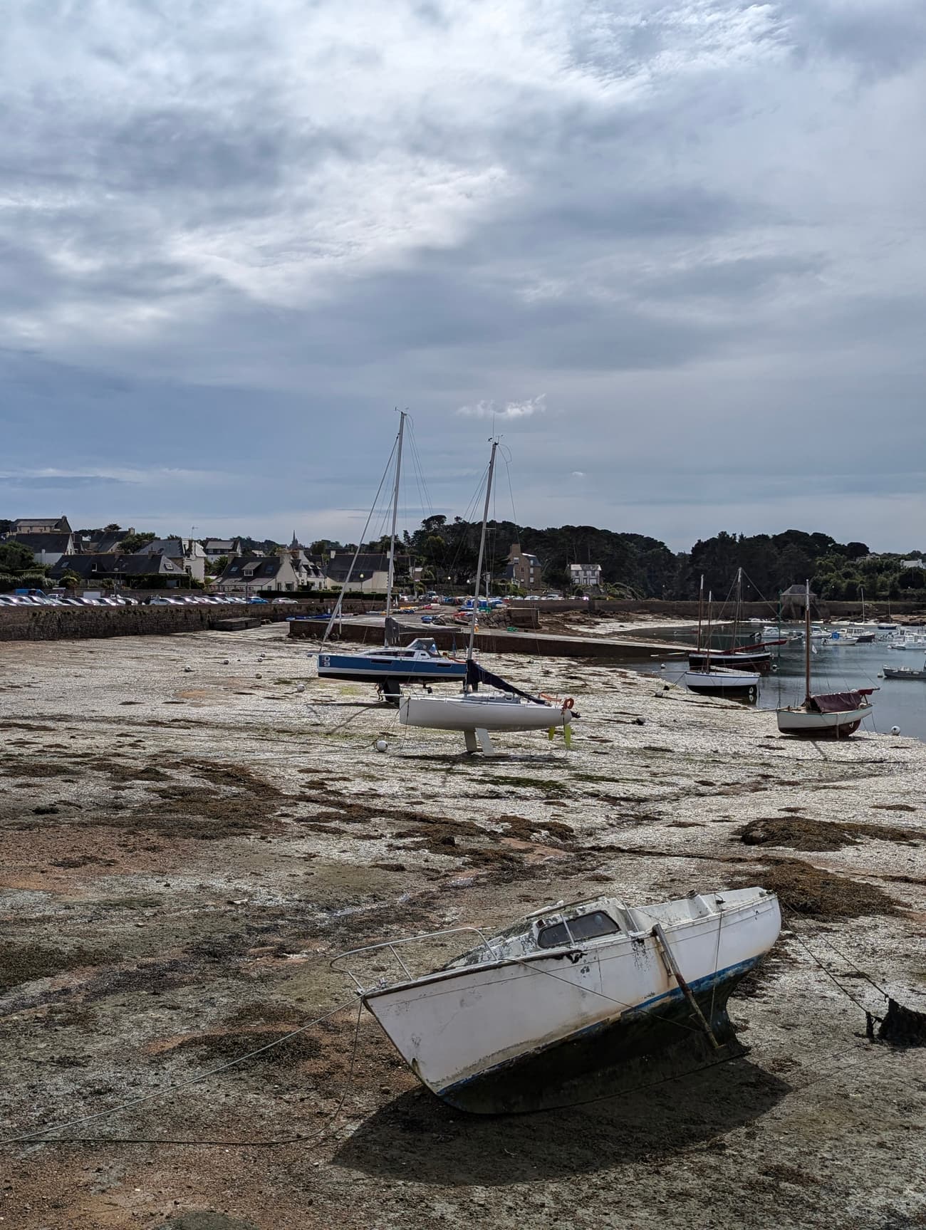 A tidal harbour drained at low water, sailing boats propped upright on the seabed and one hull tipped over in the foreground, the village behind the quay.