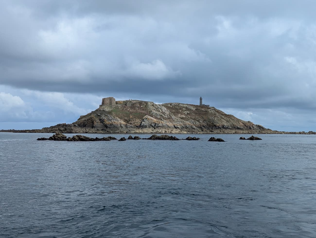 A rocky island carrying a stone fort and a lighthouse, dark reefs breaking the water in front, under heavy grey cloud.