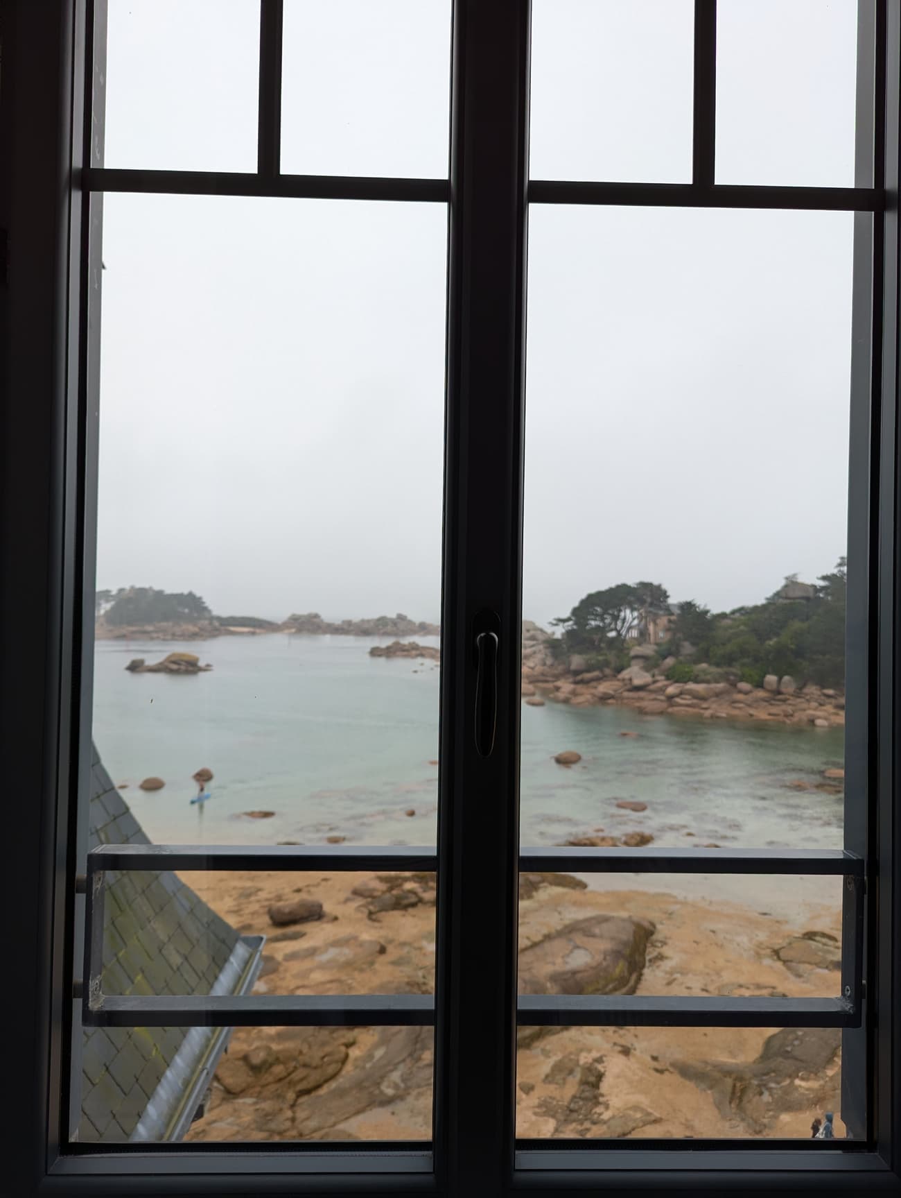 A misty pink-granite cove seen through a window: turquoise shallows, scattered boulders and a paddleboarder crossing the bay.