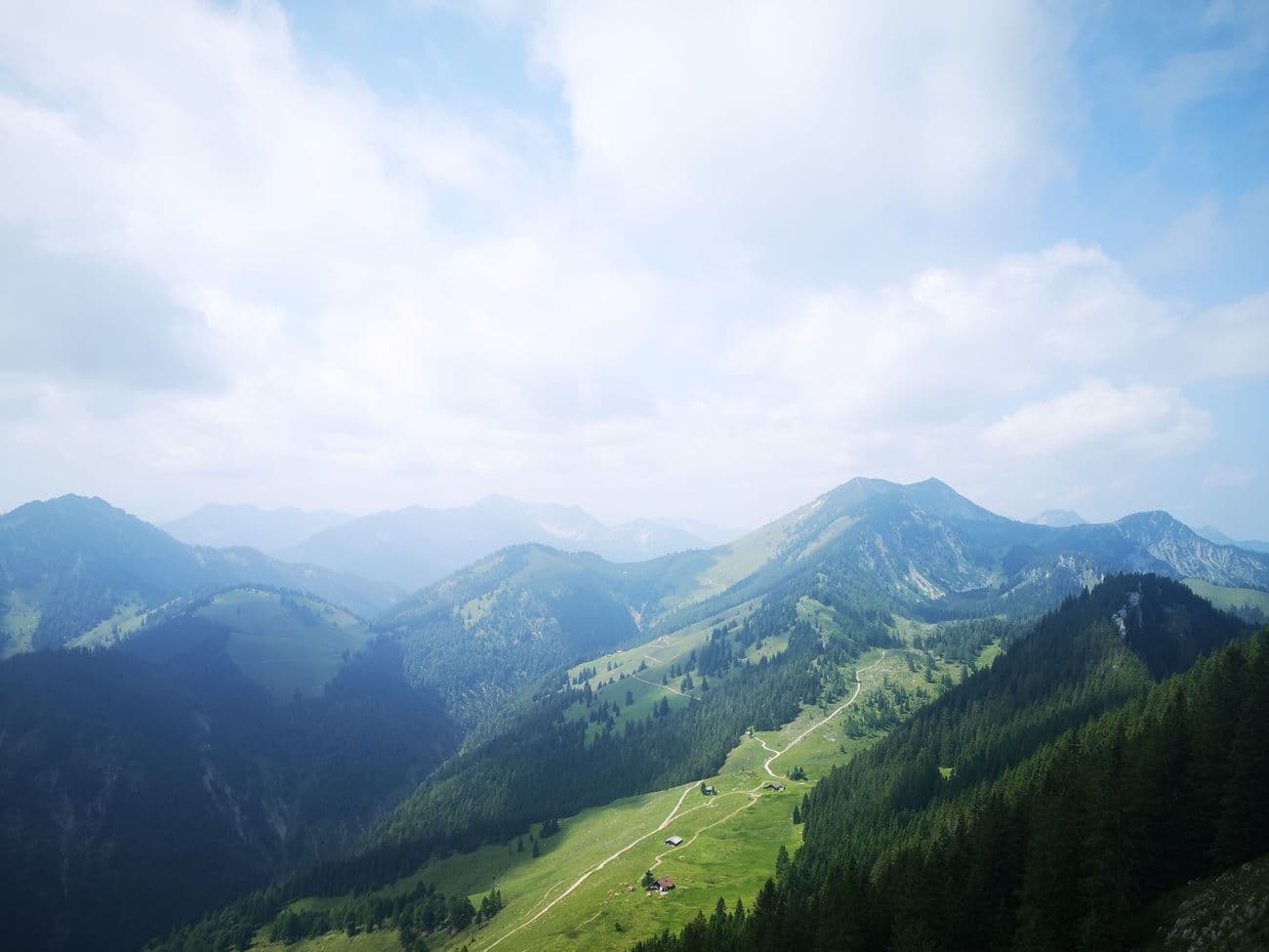 View from the Brünnstein over green alpine pastures and a gravel path, ridge after ridge fading into summer haze.