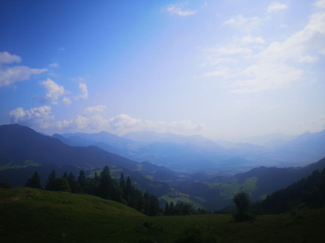 Green alpine meadow and spruce in the foreground, the wide hazy Inn valley and distant ranges beyond.