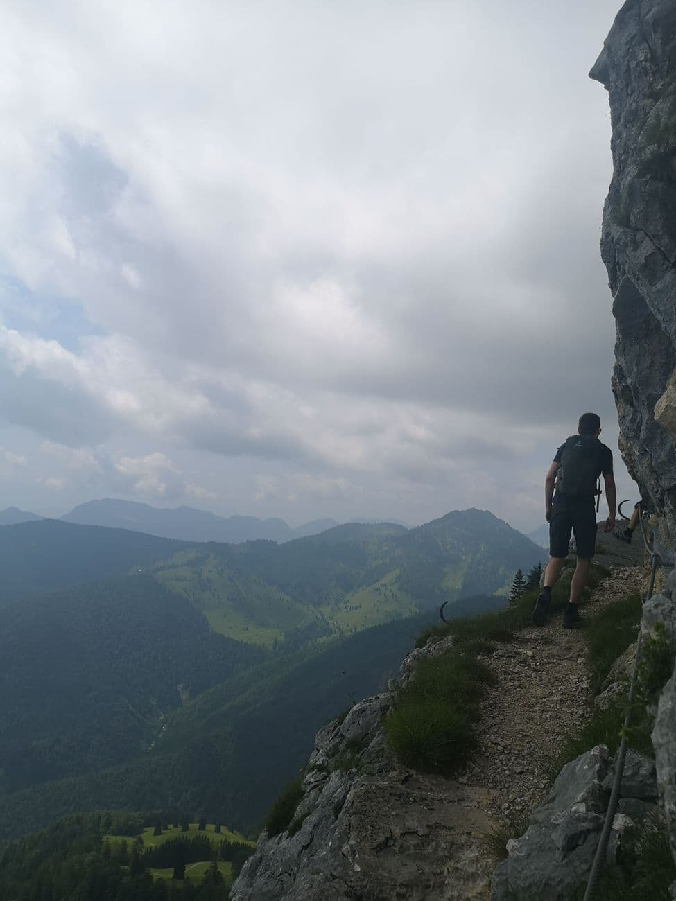 A hiker on a narrow exposed path beneath a rock overhang, safety cable to the right and the valley dropping away left.