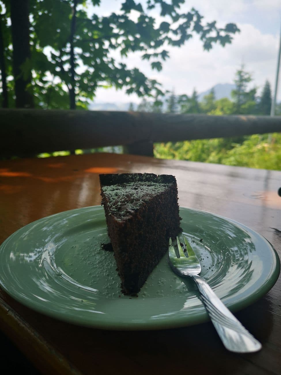 A slice of dark chocolate cake on a green plate at the Brünnsteinhaus, wet wooden table and mountains beyond the railing.