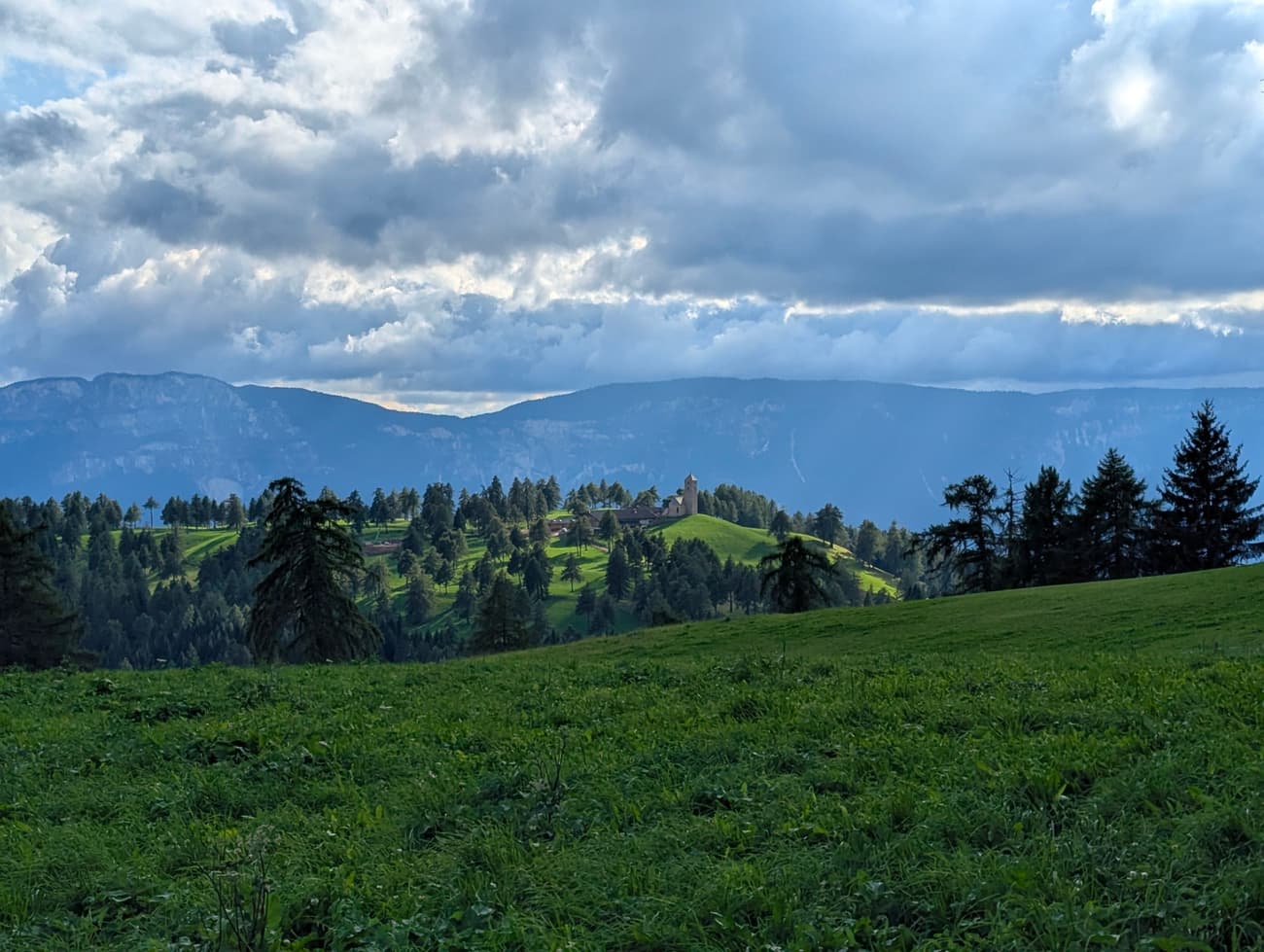 A small church on a wooded knoll above green South Tyrolean meadows, larches in the foreground and heavy cloud over the ranges behind.
