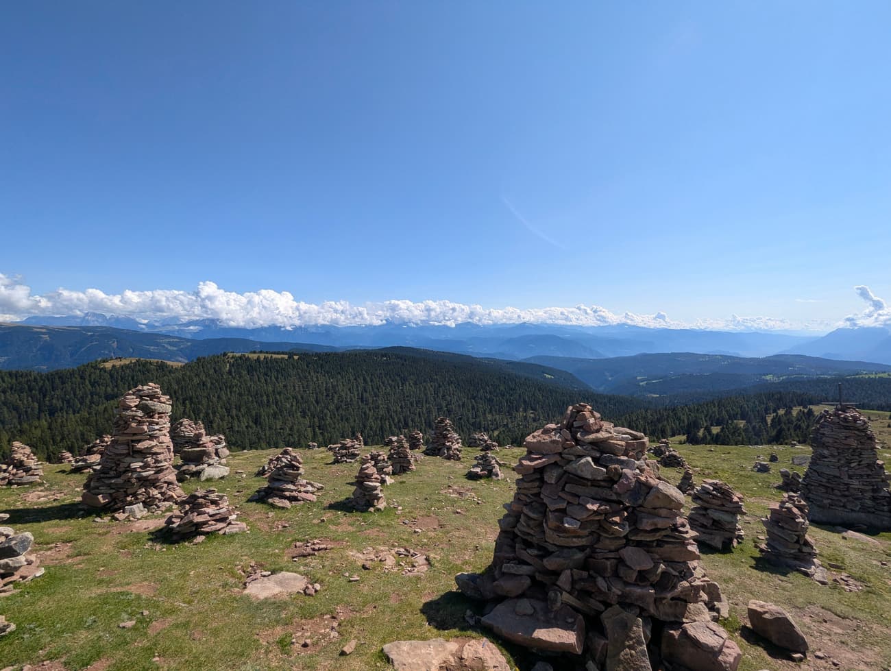 A field of stacked stone cairns on a grassy ridge, dozens of them across the slope, with the Dolomites lined up on the horizon.