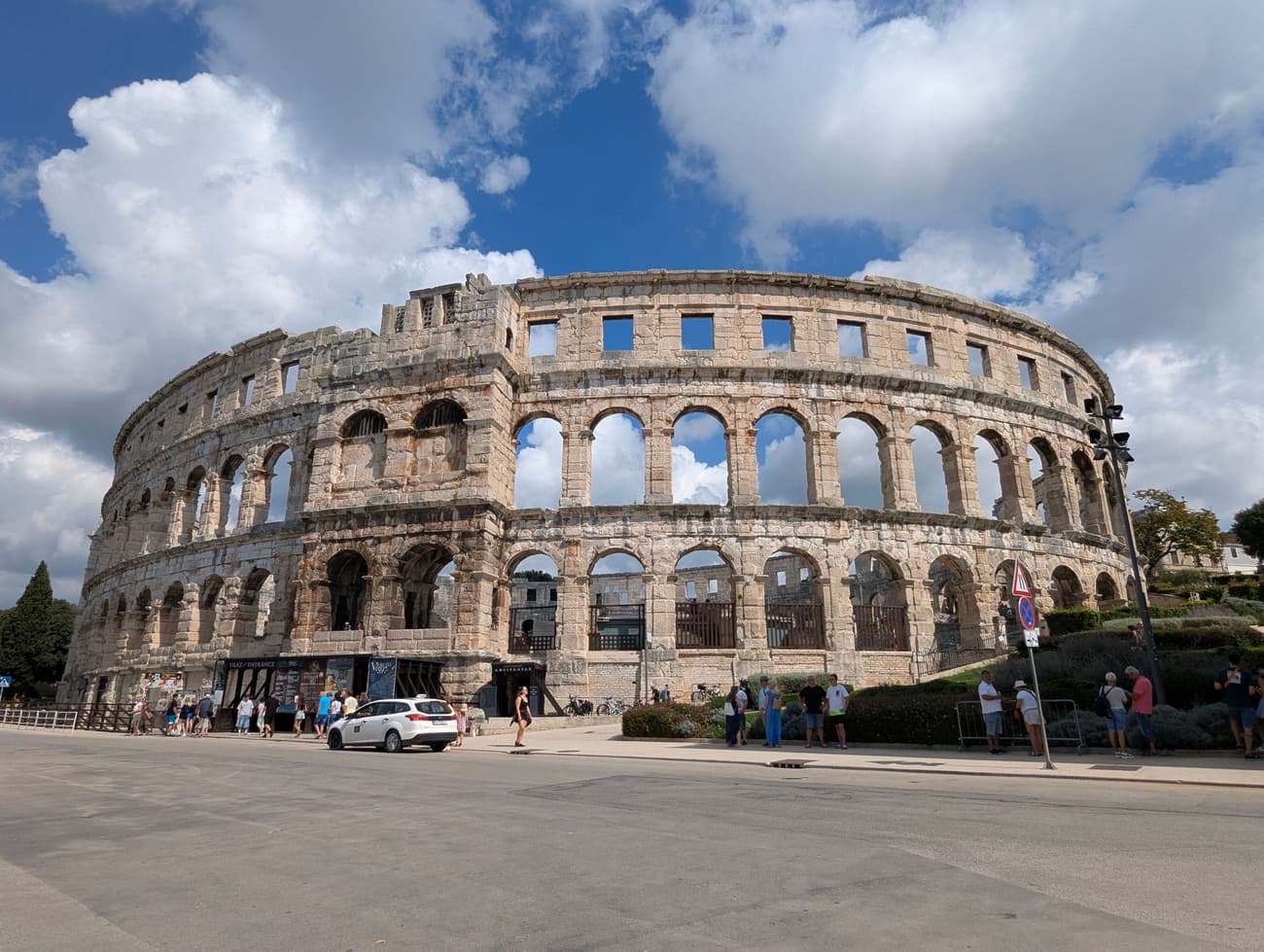 The Roman amphitheatre in Pula seen from the street: three tiers of pale limestone arches standing almost complete against towering white cloud.
