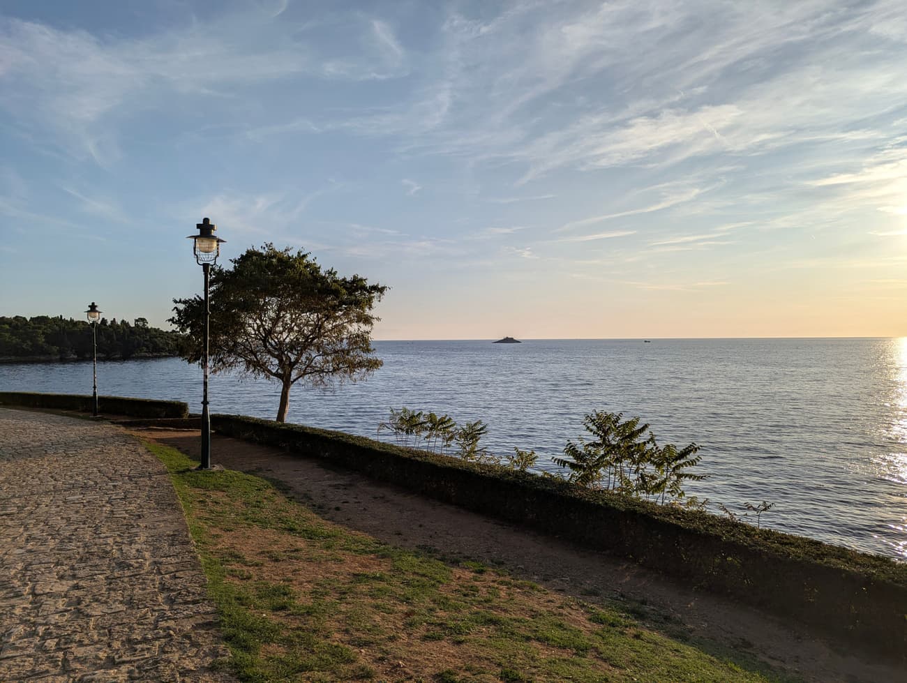 A cobbled seafront path at golden hour, a wrought-iron lamp post and a wind-shaped tree beside a low hedge, a small island out on the calm Adriatic.
