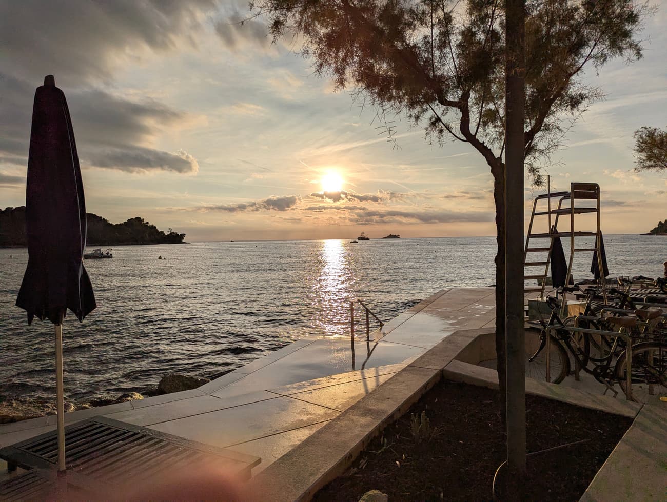 Sunset over the Adriatic from a lido: a furled umbrella, a lifeguard chair and parked bikes on the concrete apron, sun track running across the water.