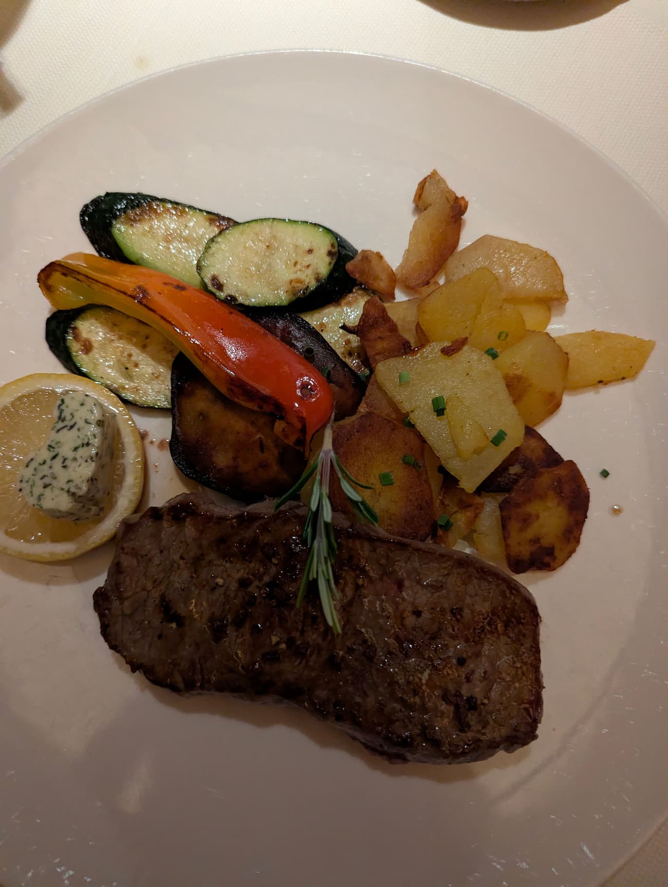 A grilled steak with a sprig of rosemary on a white plate, alongside sautéed potatoes, grilled courgette, aubergine and peppers, and herb butter on lemon.