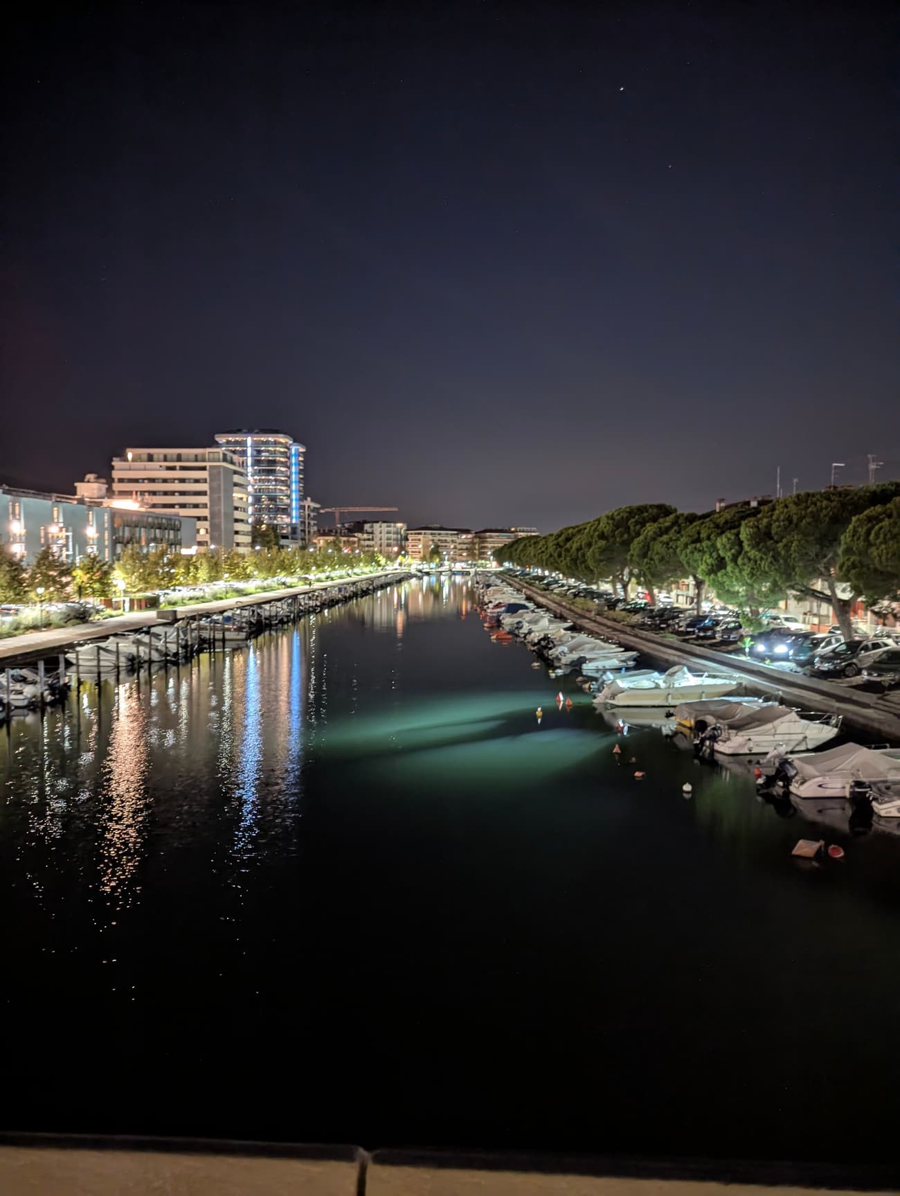 A marina canal at night: motorboats moored along both quays, umbrella pines on the right, hotel lights streaking across the black water.