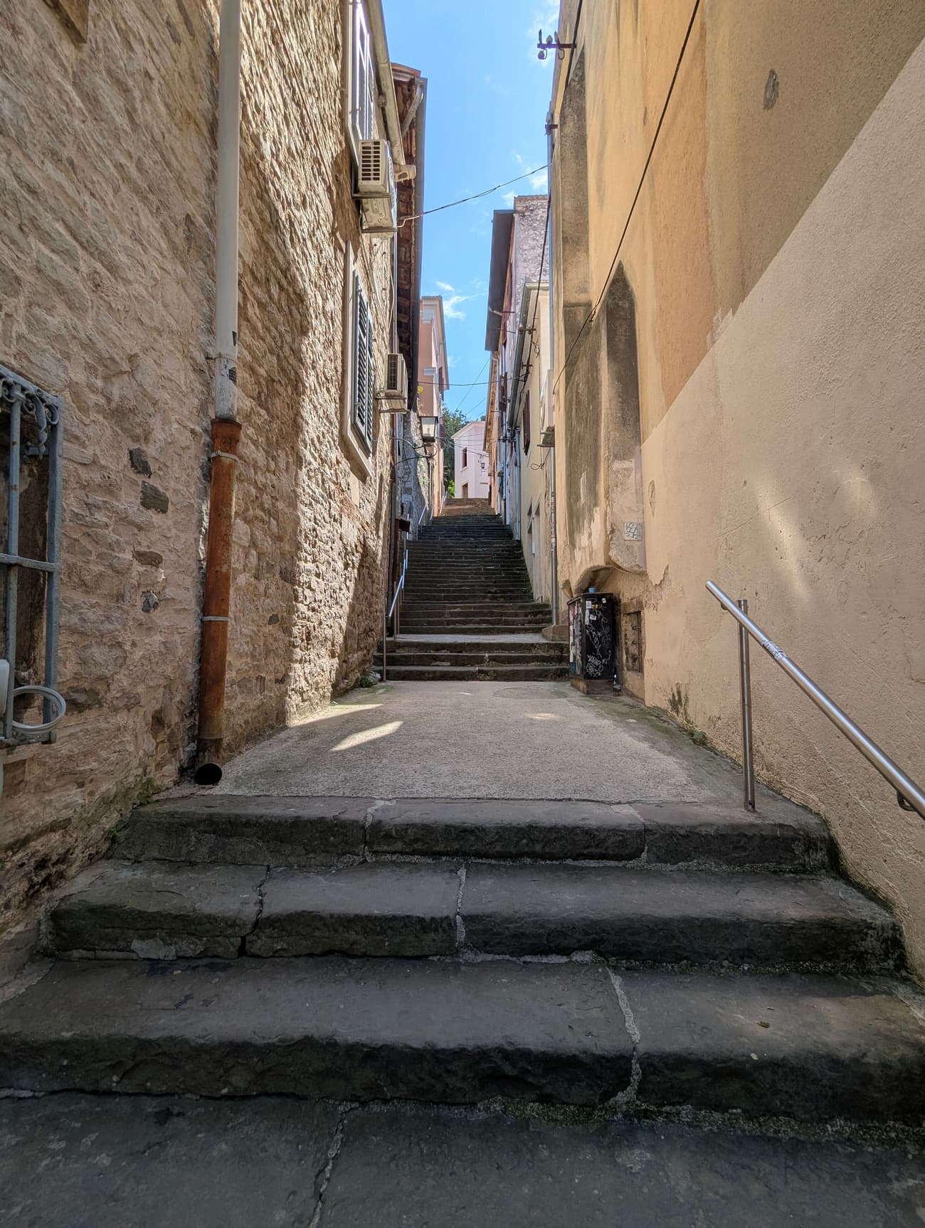 A narrow stepped alley climbing between stone and ochre-plastered houses in a Croatian old town, a strip of blue sky overhead.