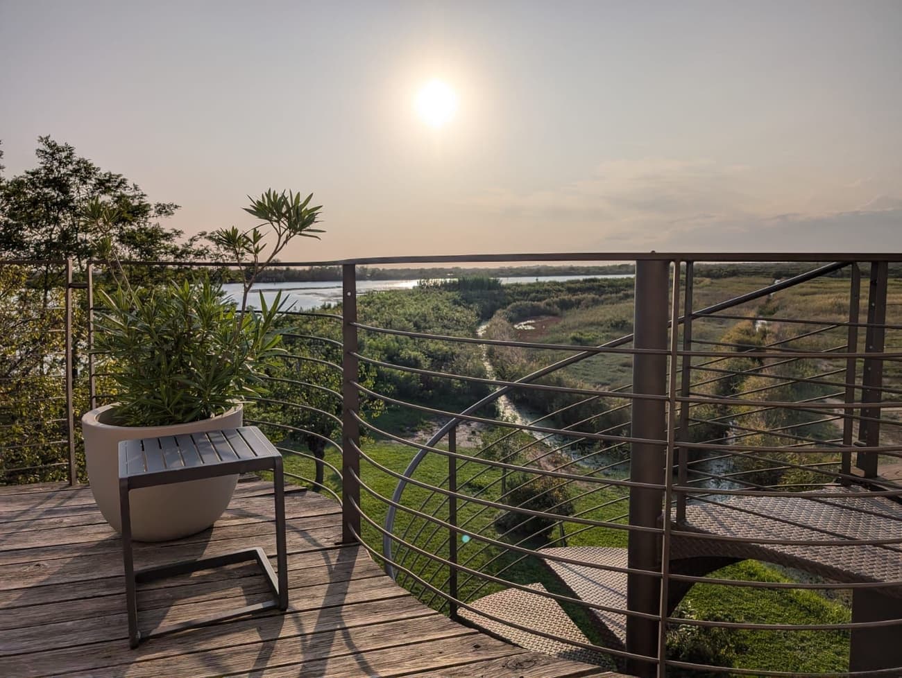 A wooden terrace with a curved steel railing at sunset, a potted oleander in the corner and reed beds and channels stretching to the lagoon.