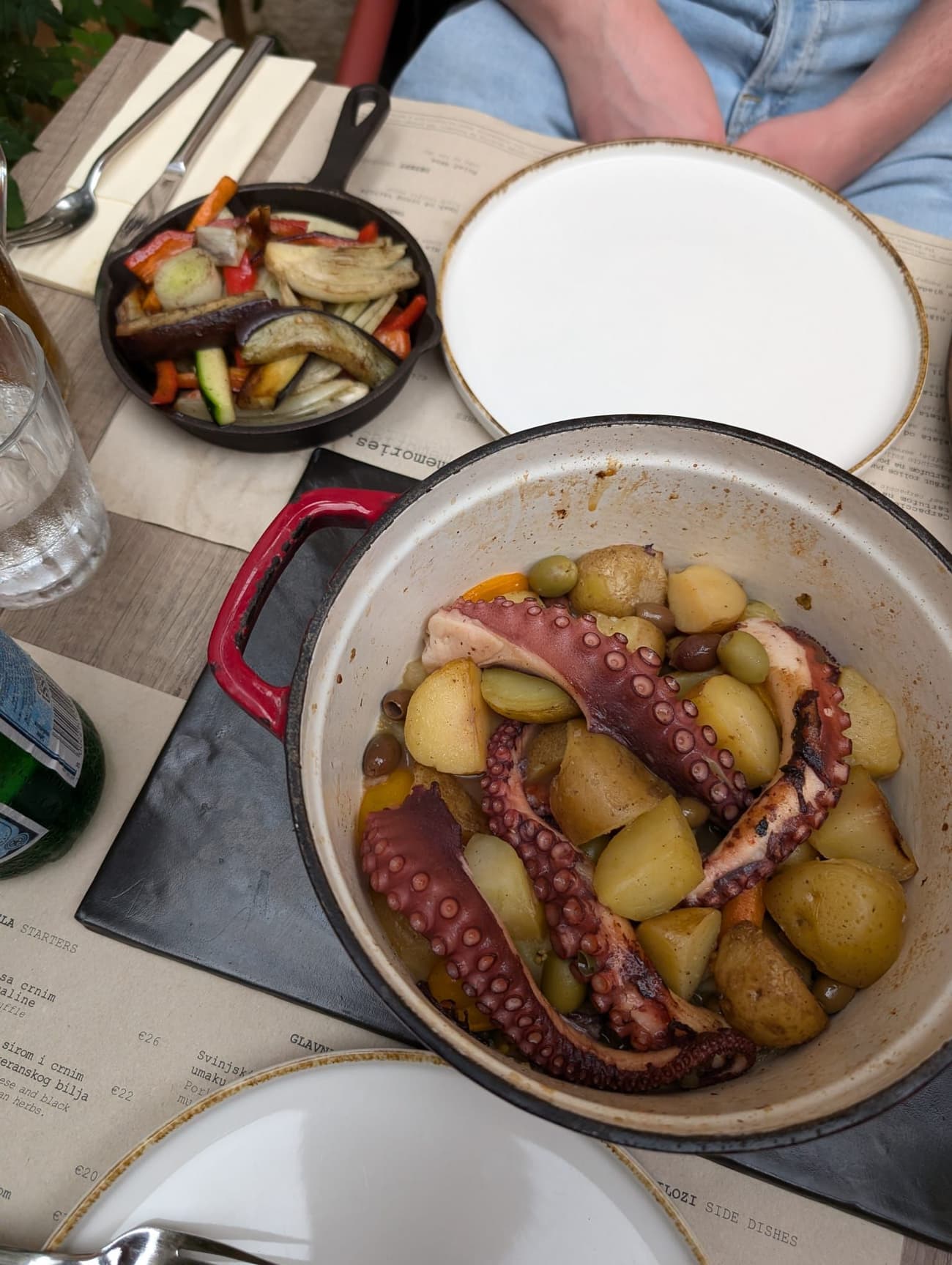 Octopus slow-cooked with potatoes and olives in a cast-iron pot, a skillet of grilled peppers, fennel and aubergine alongside on a restaurant table.