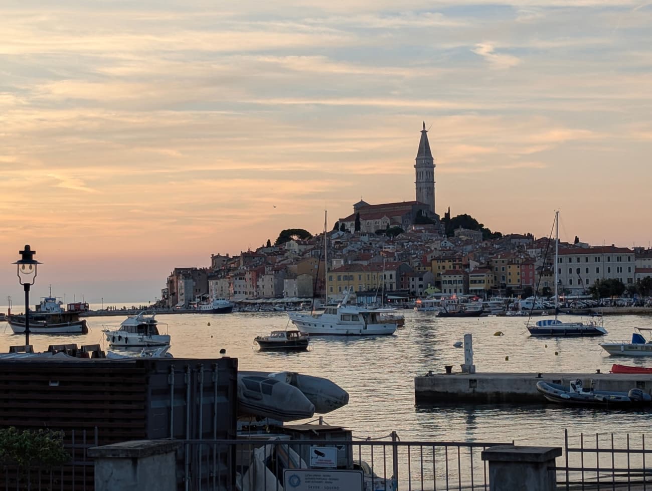 Rovinj at sunset from across the harbour: the bell tower of St Euphemia above tiers of ochre and terracotta houses, boats moored on still pink water.