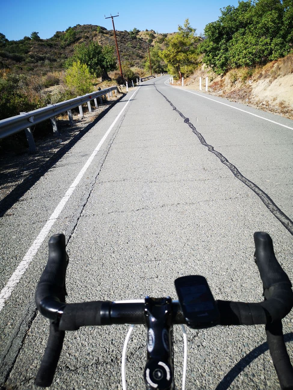 Handlebar view from a road bike with a GPS computer, an empty patched road running into dry Cypriot hills.