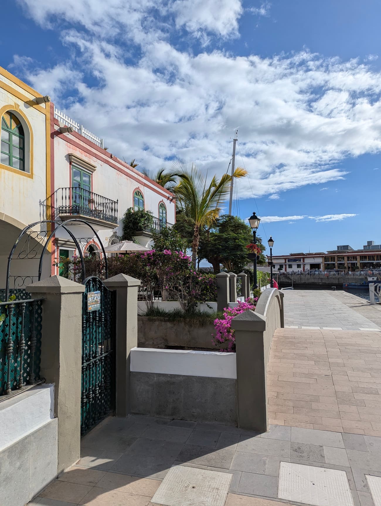 The harbour promenade at Puerto de Mogán: pastel houses with green shutters and wrought-iron balconies, bougainvillea and palms, a mast rising behind.
