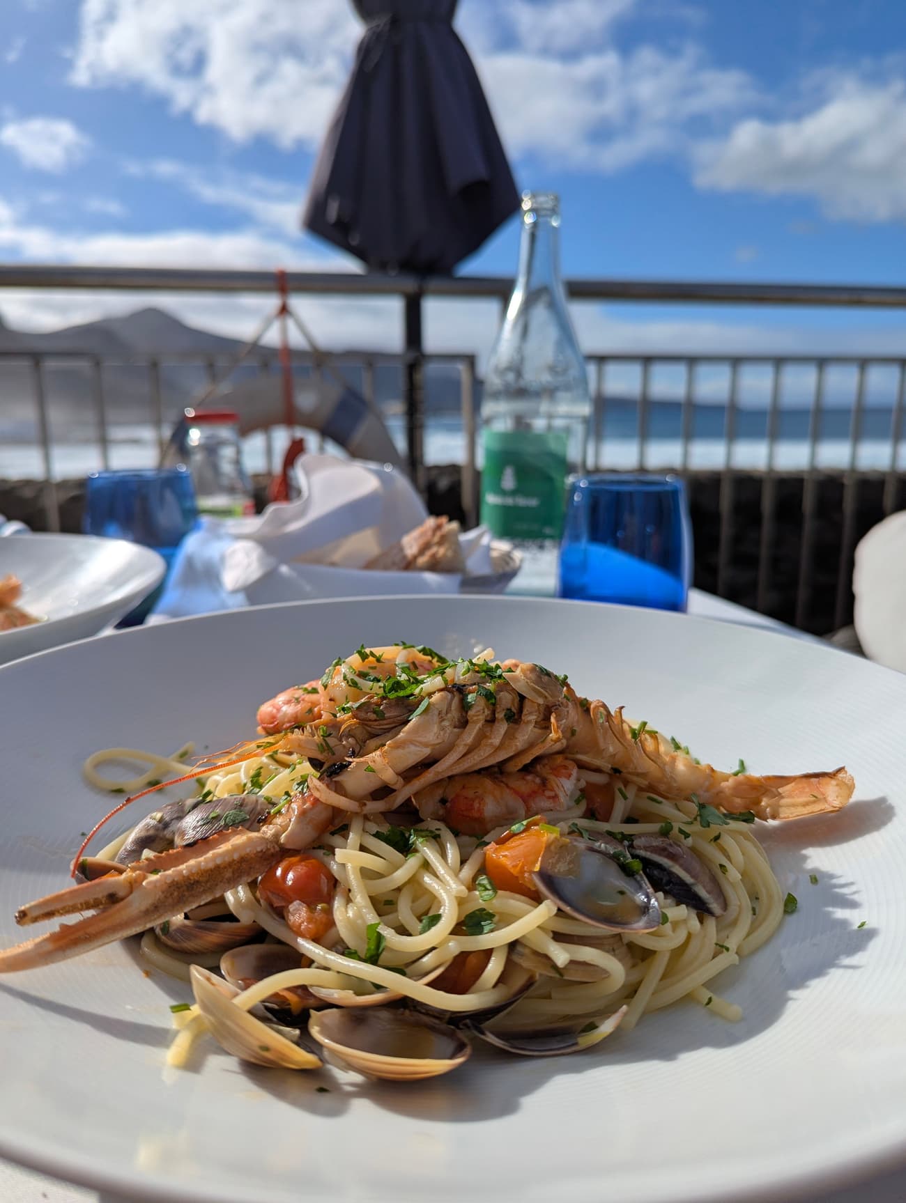 Seafood spaghetti with langoustine, prawns and clams on a terrace table, the sea and a lifebuoy just beyond the railing.