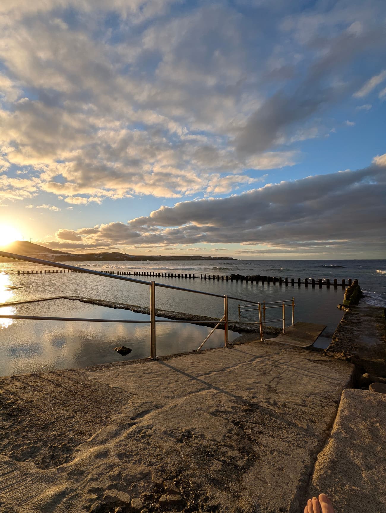 A seawater tidal pool at sunset, handrails leading down to the water and the low sun breaking through cloud over the coast.