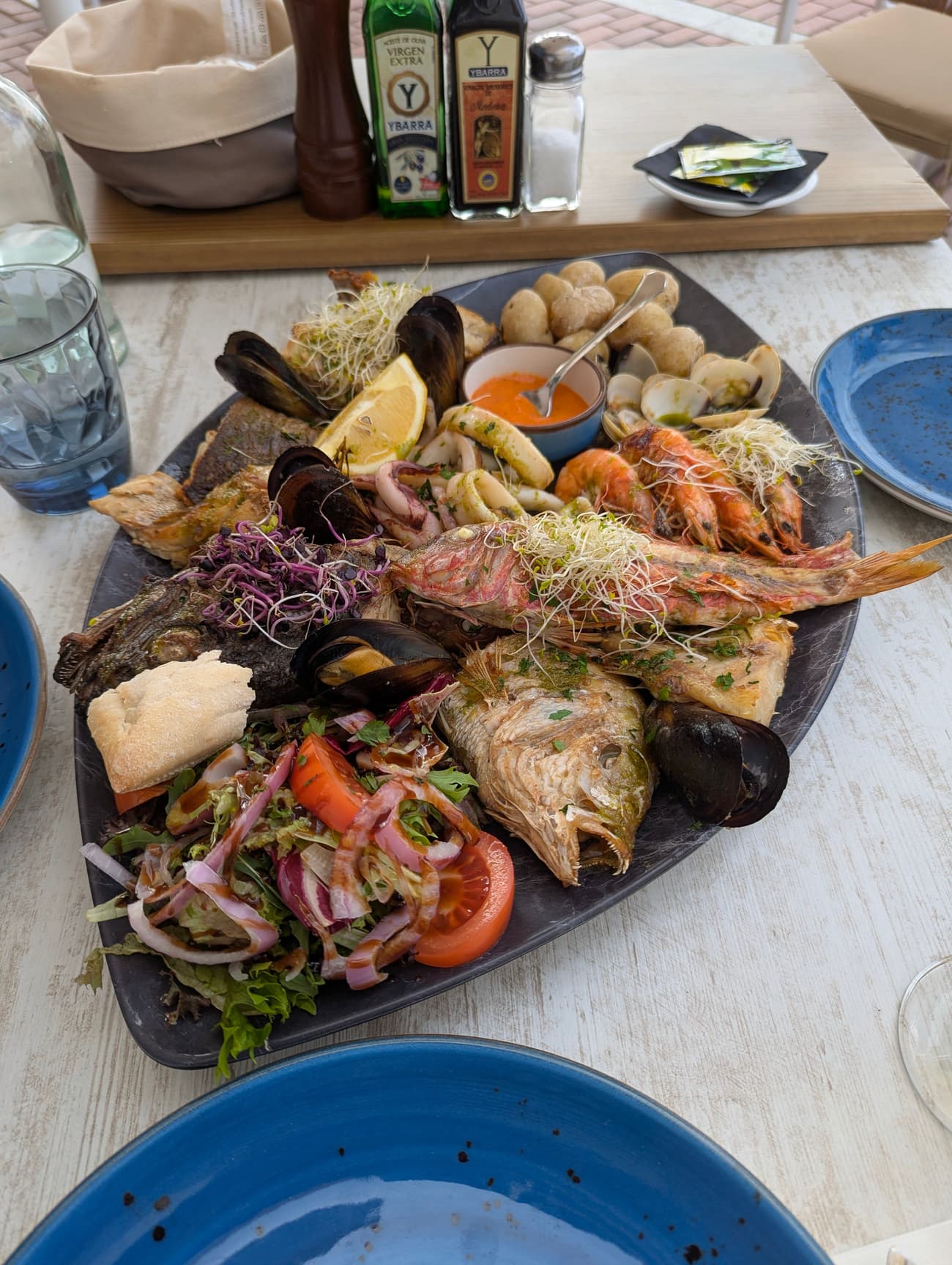 A large seafood platter — whole grilled fish, mussels, clams, prawns, squid and papas arrugadas — on a slate dish with salad and bread.