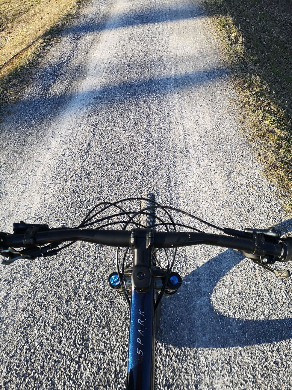 Handlebar view along a gravel farm track, the blue Scott Spark frame and the rider's long shadow stretching ahead in low sun.