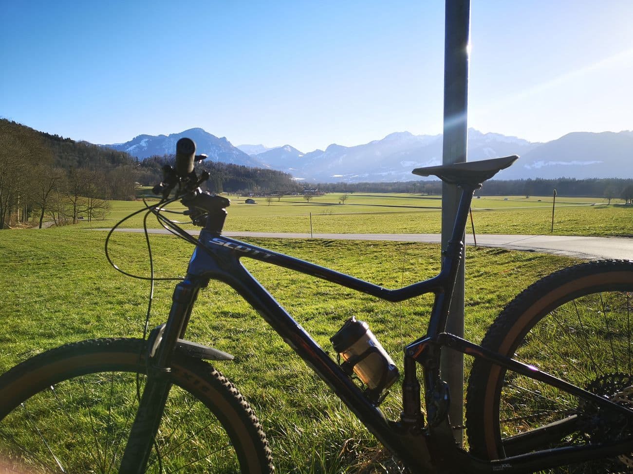A blue Scott Spark mountain bike leaning against a post in a green meadow, the snow-dusted Alps on the horizon.