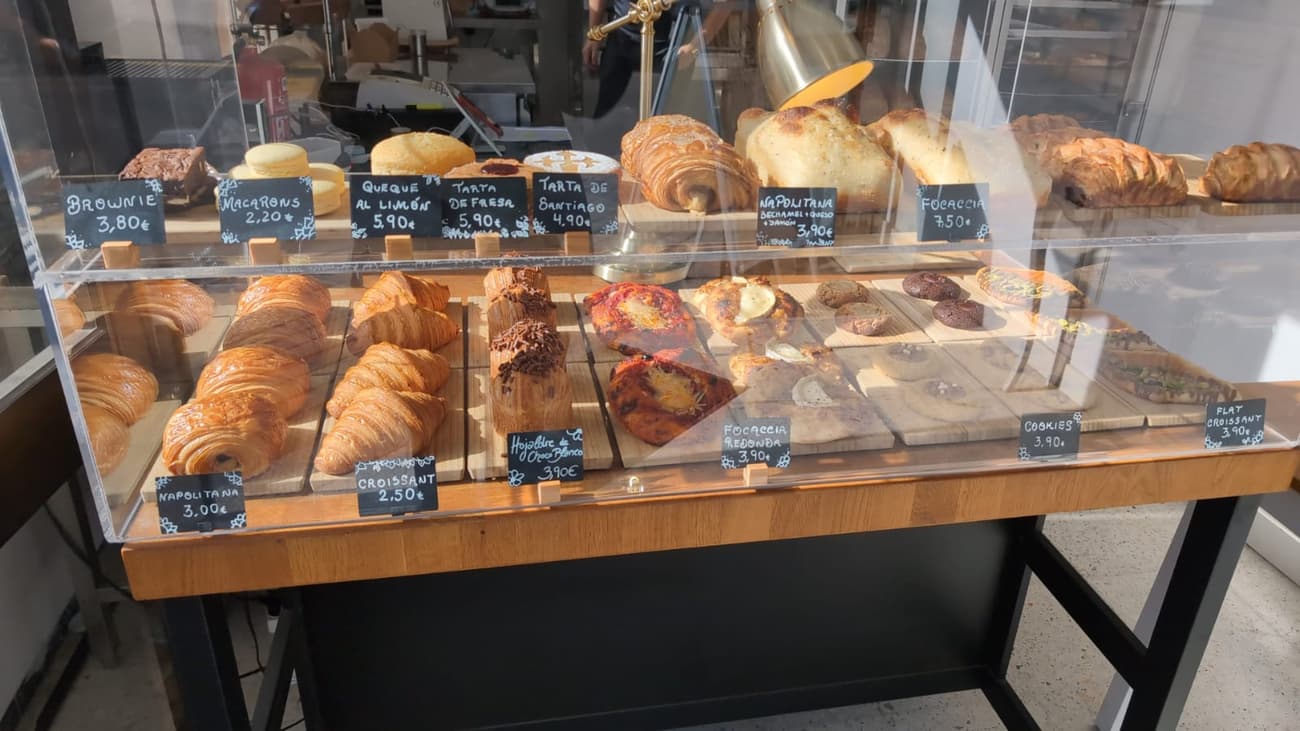 A bakery counter behind glass: rows of croissants, napolitanas and focaccia on wooden boards with hand-chalked price cards in Spanish.