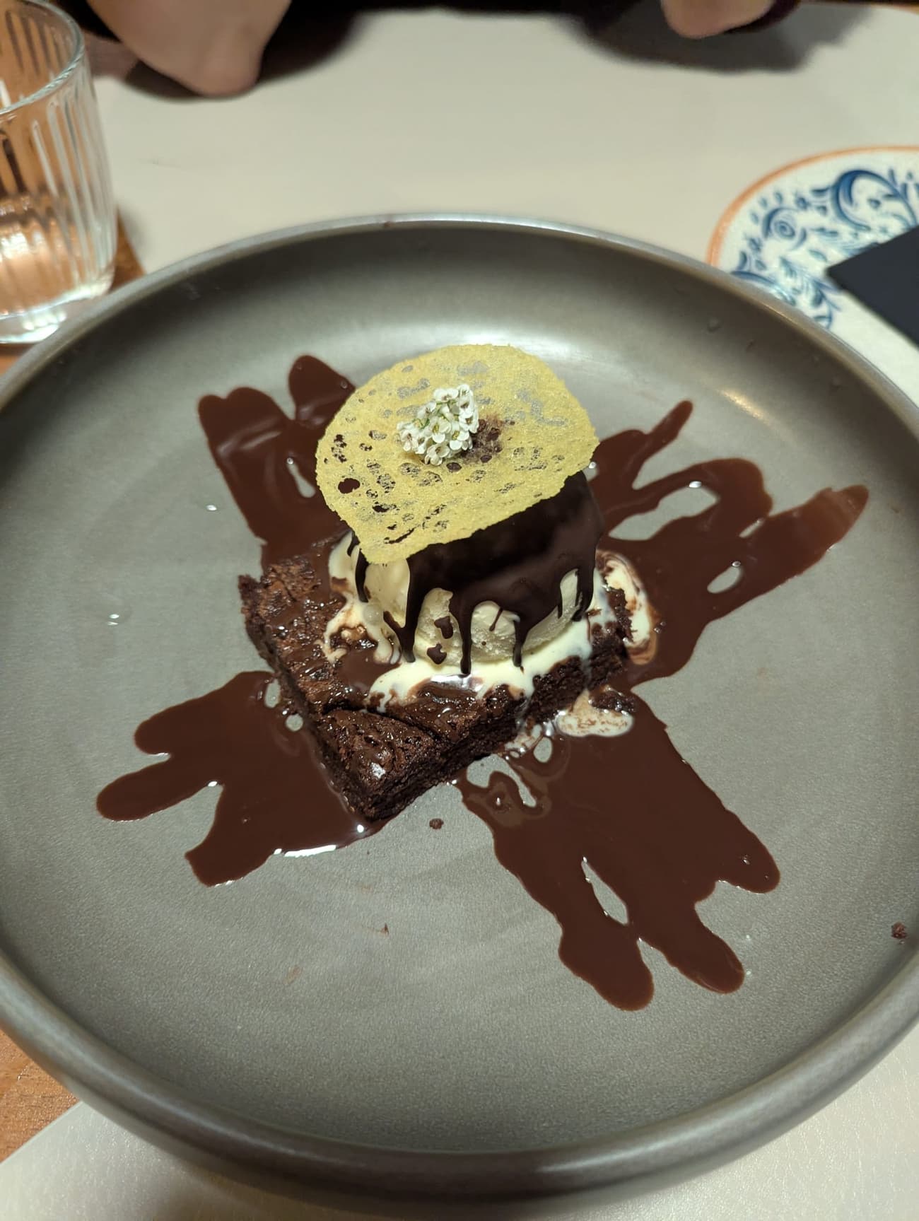 A chocolate brownie with ice cream and dark sauce on a grey plate, finished with a crisp tuile and a white flower.