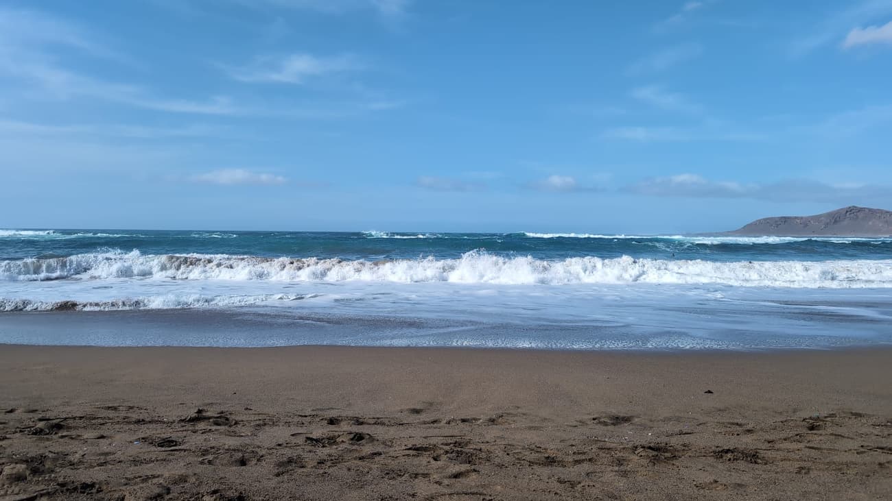 Waves rolling onto a wide sandy beach under a bright blue sky, a low volcanic headland closing the bay to the right.