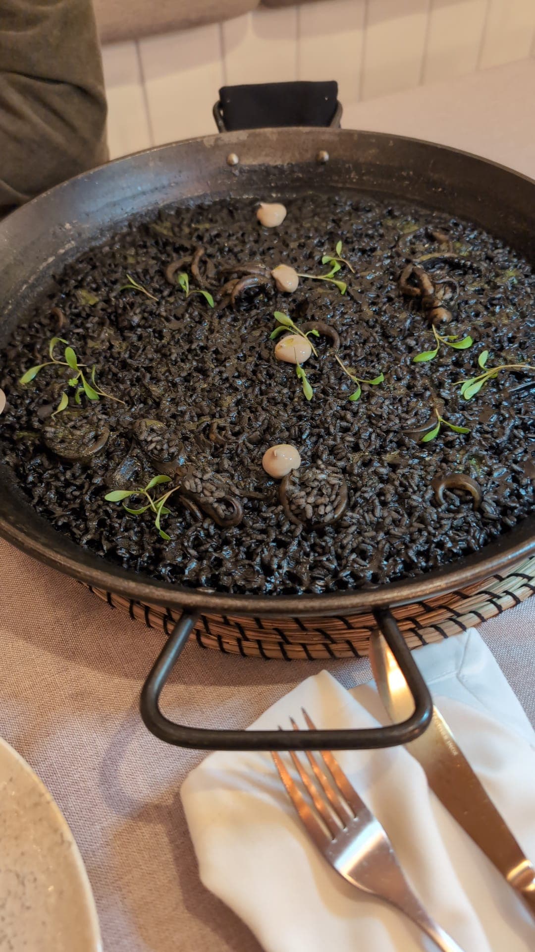 Arroz negro served in a steel paella pan: squid-ink rice with rings of squid, dots of aioli and small green herb leaves.