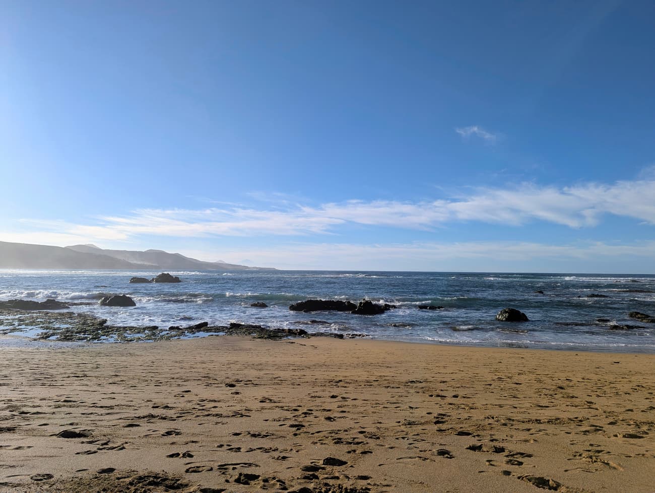 A wide sandy beach at low tide with a dark volcanic reef exposed offshore, the coast running north and Teide faint on the horizon.