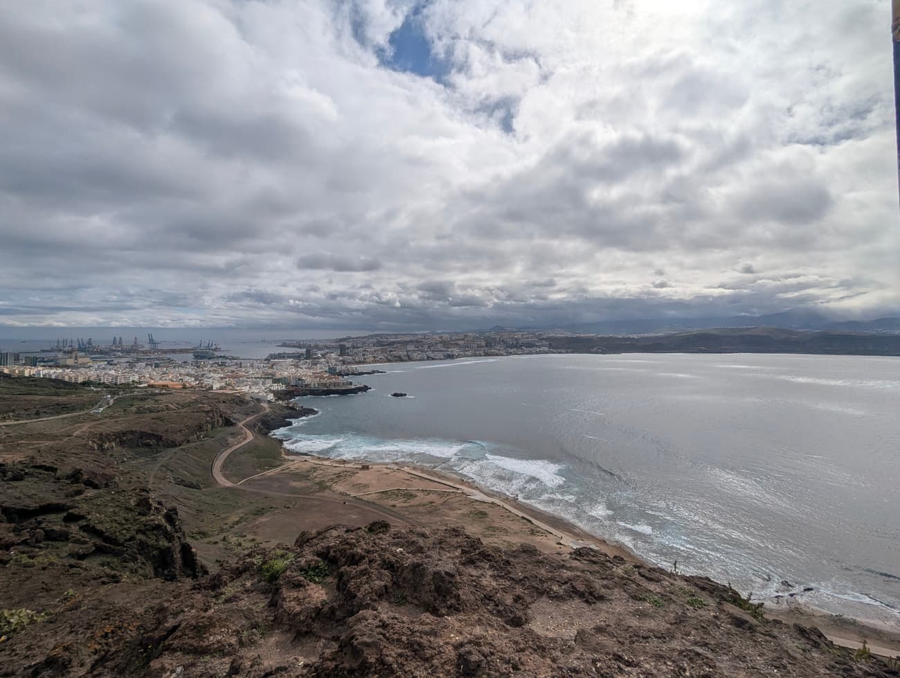 Las Palmas from the clifftop above La Isleta: the bay curving round to the port cranes and the city spread along the shore under heavy cloud.