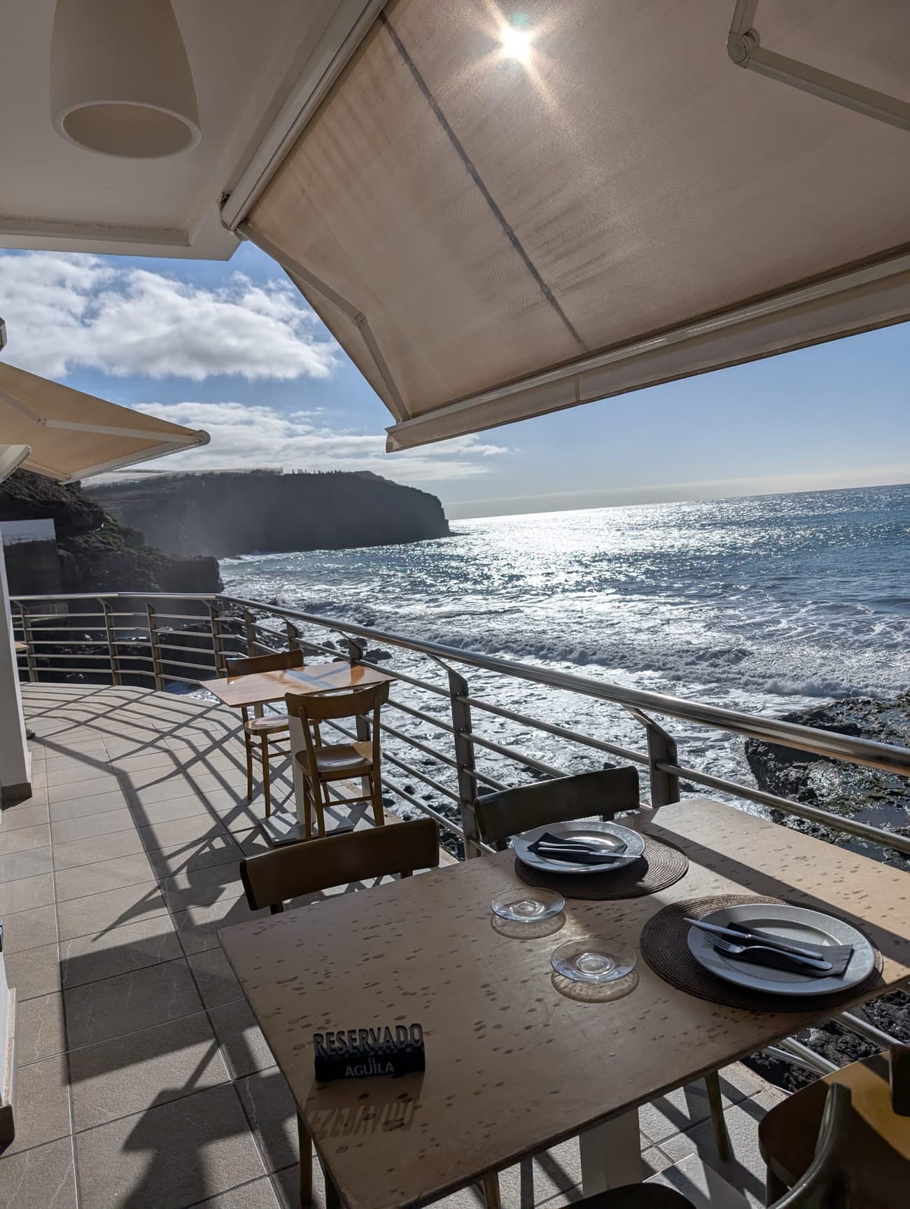 A restaurant terrace under a cream awning above a rocky shore, tables laid and a 'Reservado' card out, surf breaking on black volcanic rock below.