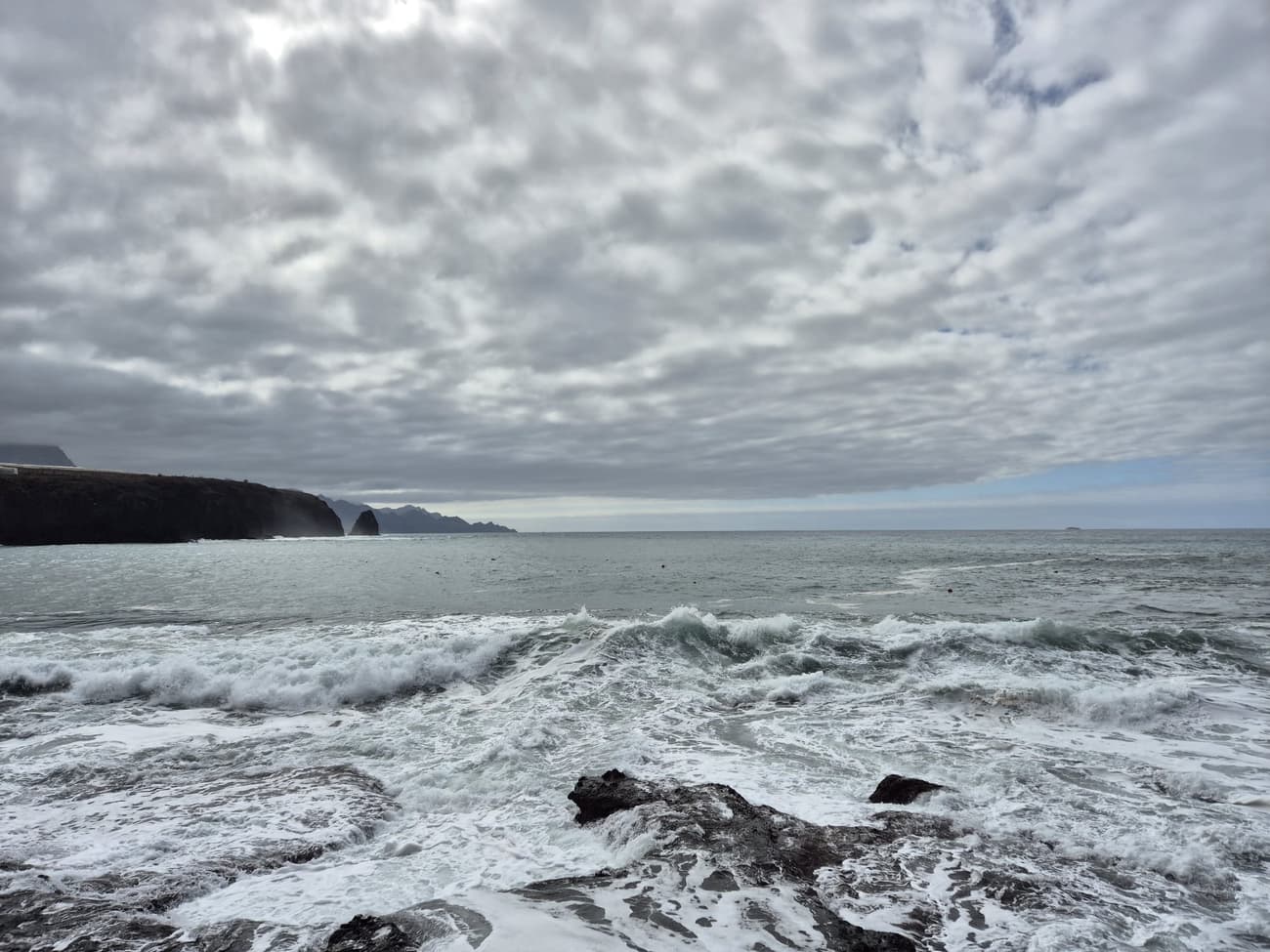 Heavy surf breaking over black volcanic rock on the north coast, dark cliffs running out to a sea stack under a thick mackerel sky.