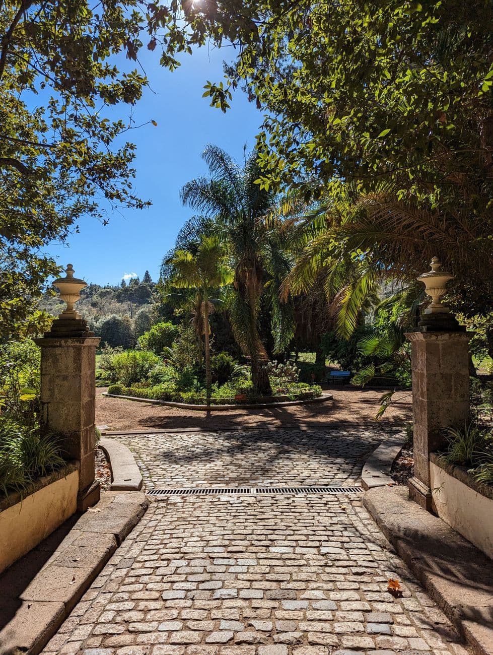 A cobbled path running between stone gateposts topped with urns into a garden of palms and flowerbeds.
