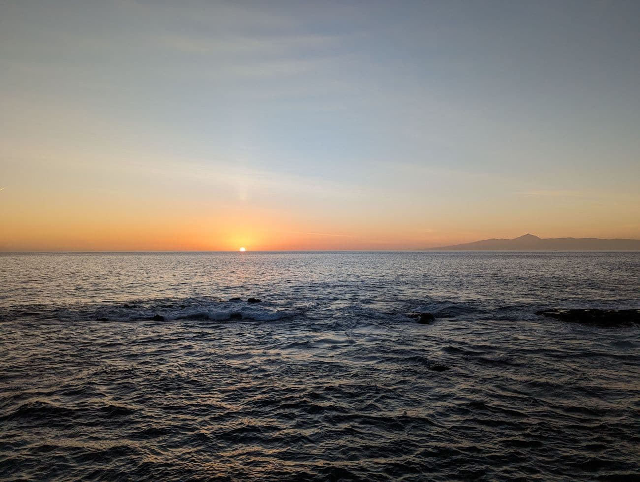 Sunset over the Atlantic from a rocky shore, the cone of Teide on Tenerife just visible across the water.