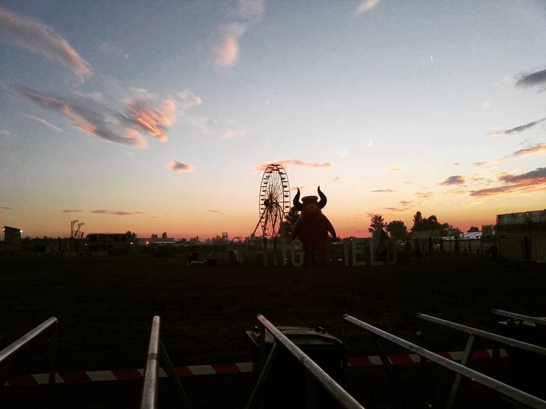 Highfield Festival at dusk: a ferris wheel and a large horned mascot silhouetted against an orange sunset, seen across empty crowd barriers.