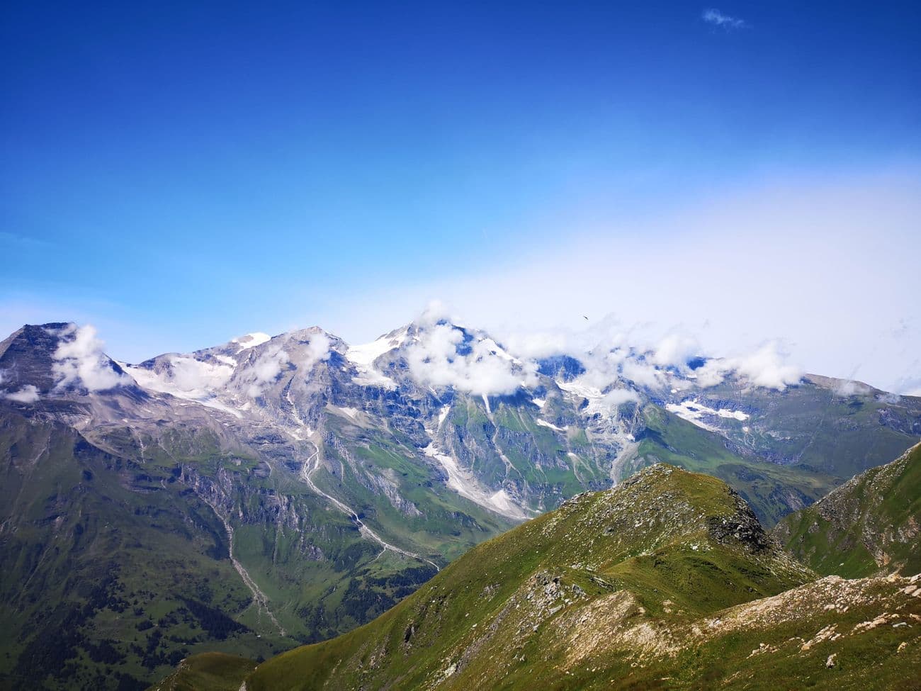 Cloud catching on a line of glaciated peaks above green alpine slopes, seen from a pass road under deep blue sky.