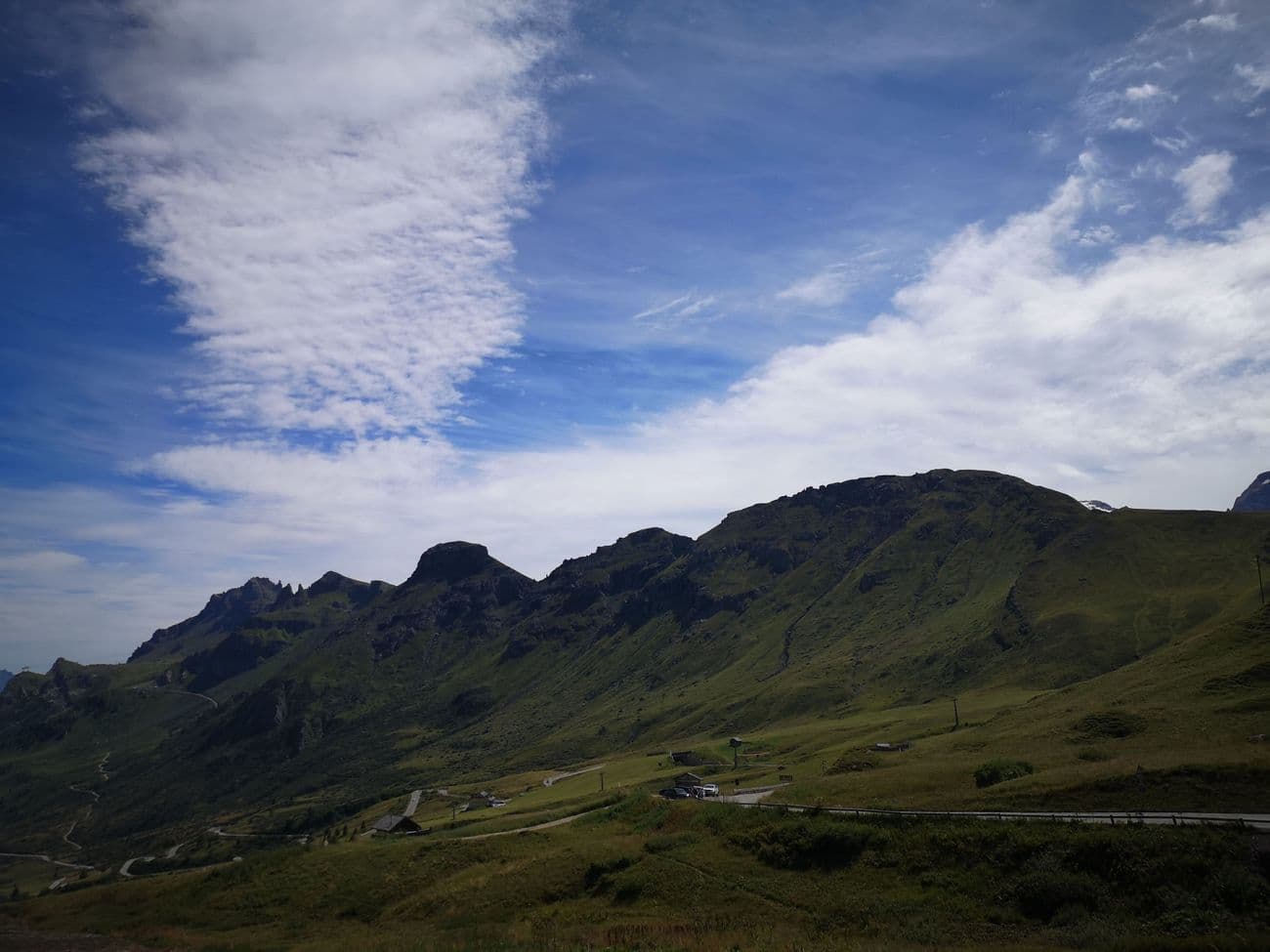 A grassy alpine pass with a rocky crest above, the road curling past a handful of huts and a parking area.