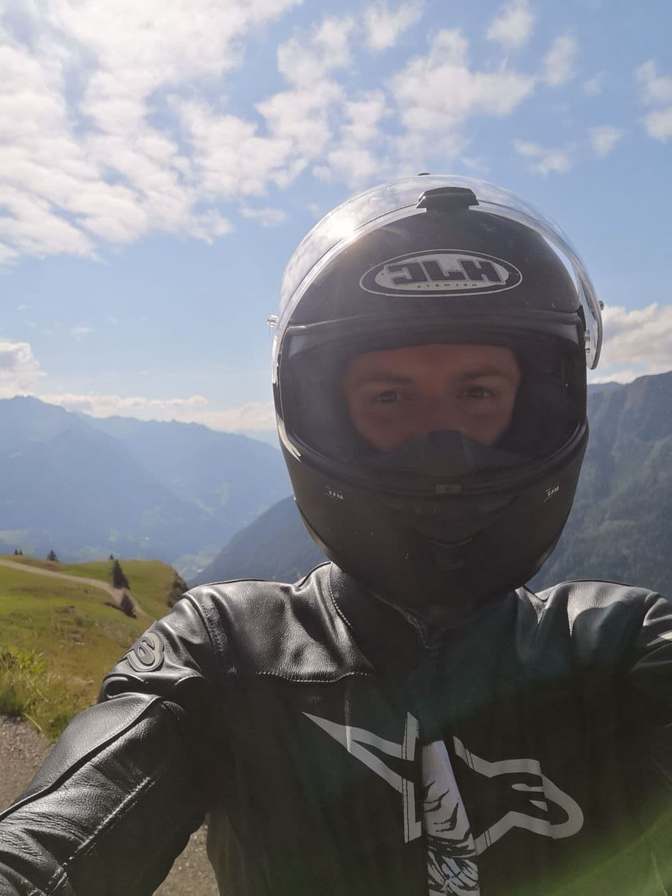 A selfie in a full-face helmet and black leather suit at the roadside, alpine ridges and a green valley falling away behind.