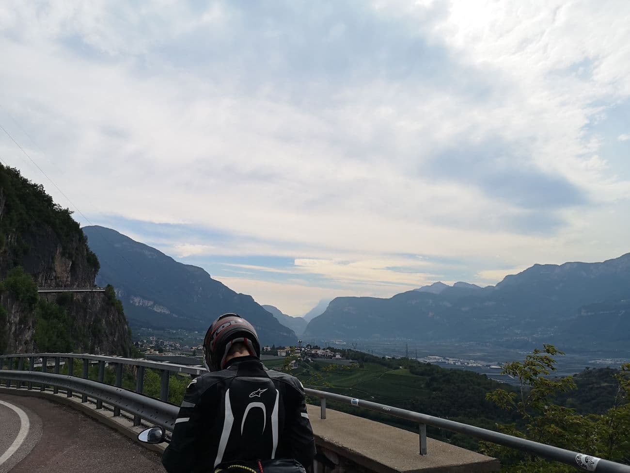 A motorcyclist at a guardrail viewpoint looking down the Adige valley, vineyards and villages below and mountains closing in on both sides.