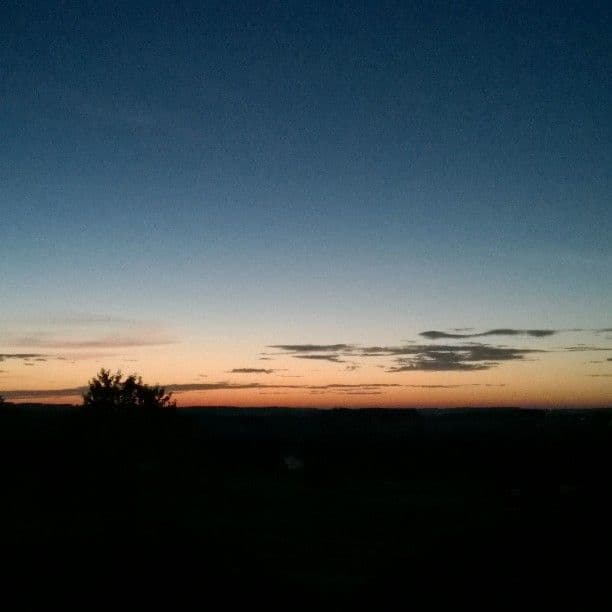 A summer sunset over flat countryside, thin bands of cloud lit orange above a dark treeline.